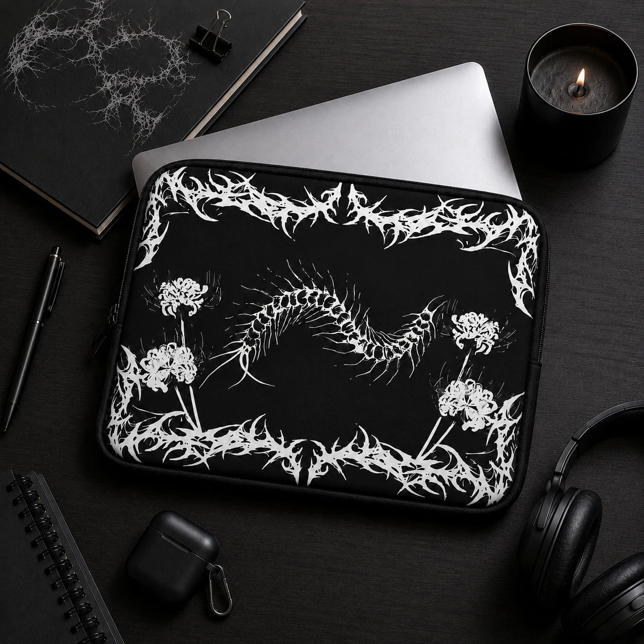 Gothic centipede laptop sleeve featuring spider lily flowers and black metal inspired artwork, displayed in a dark aesthetic workspace with laptop, headphones and black accessories.