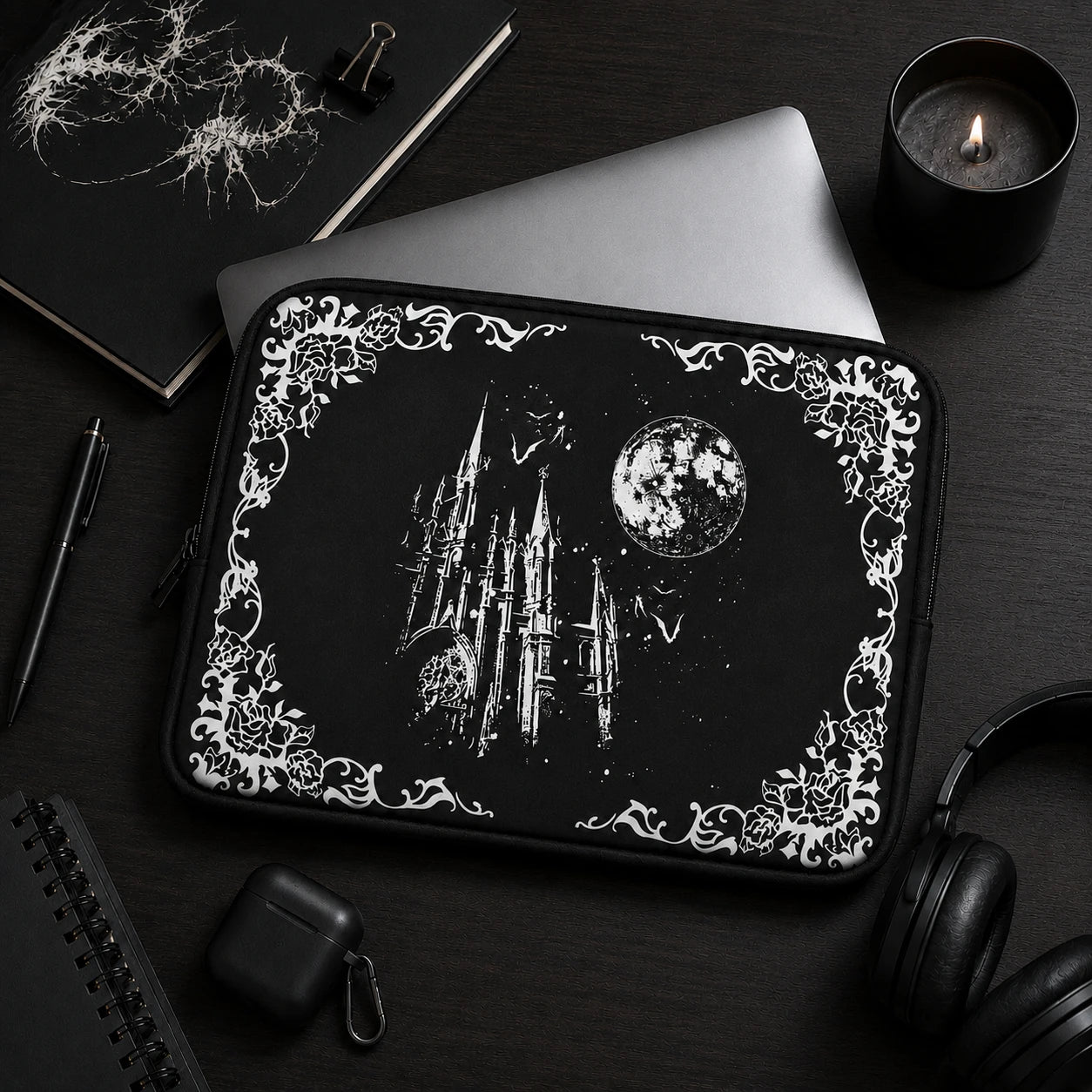 Black gothic laptop sleeve featuring a moonlit castle, bats and floral gothic border, styled in a dark academia workspace with notebook, candle and headphones, ideal for alternative and gothic aesthetics.