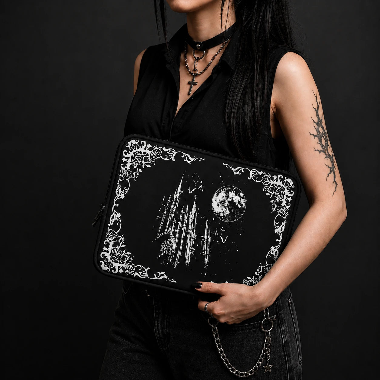 Alternative gothic woman holding a black laptop sleeve with moonlit castle, bats and ornate floral frame design, styled in a dark fashion setting with gothic accessories and black clothing.
