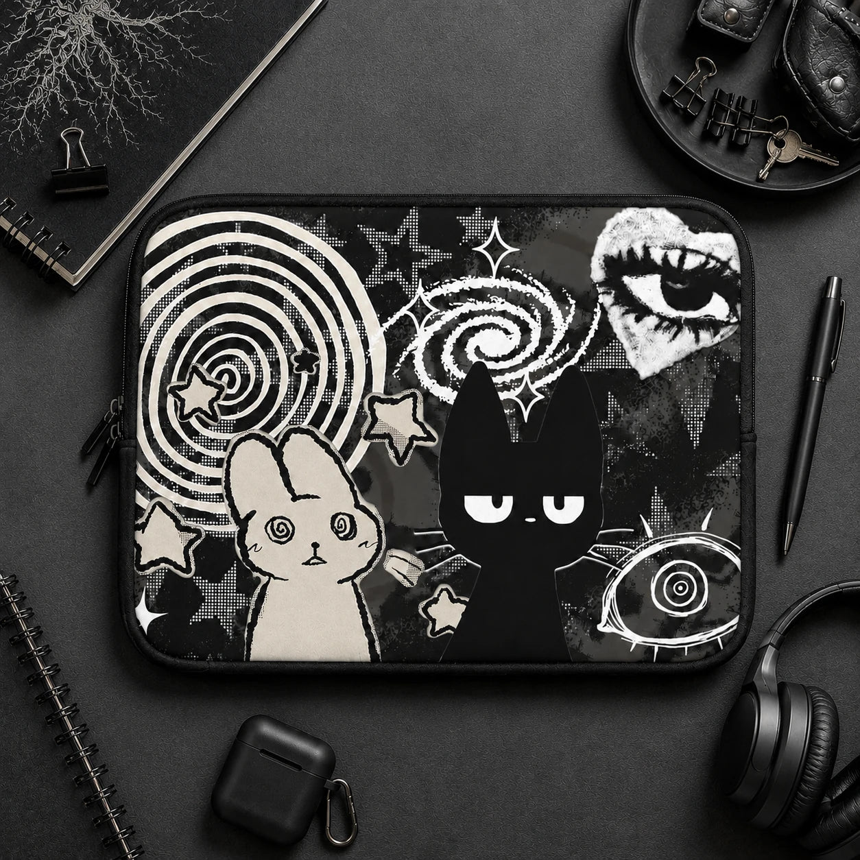Gothic aesthetic laptop sleeve with black cat and hypnotic bunny illustration displayed on a dark desk setup with headphones, notebook and alternative lifestyle accessories. 