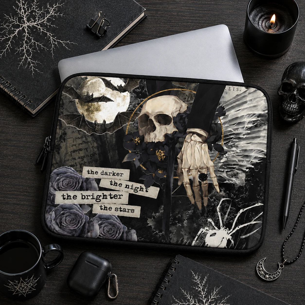 Dark gothic laptop sleeve with skull, bats, black roses and skeletal hand illustration displayed in a moody black desk setup with candles, notebooks, coffee mug and alternative lifestyle accessories.
