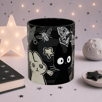 Close-up of gothic kawaii cat mug featuring weirdcore artwork, spiral stars, moths, and black celestial design.